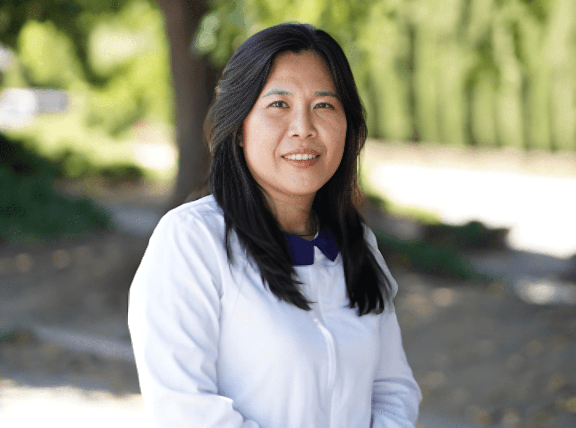 Person with long dark hair wearing a white shirt, standing outdoors with greenery in the background.
